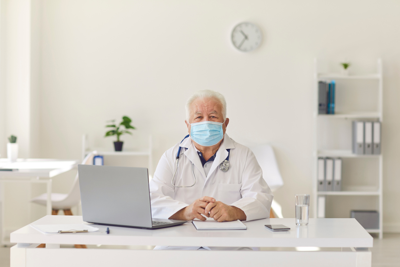 Portrait of a Senior Doctor in Protective Mask Who Sits with His Desk and Looking at the Camera.